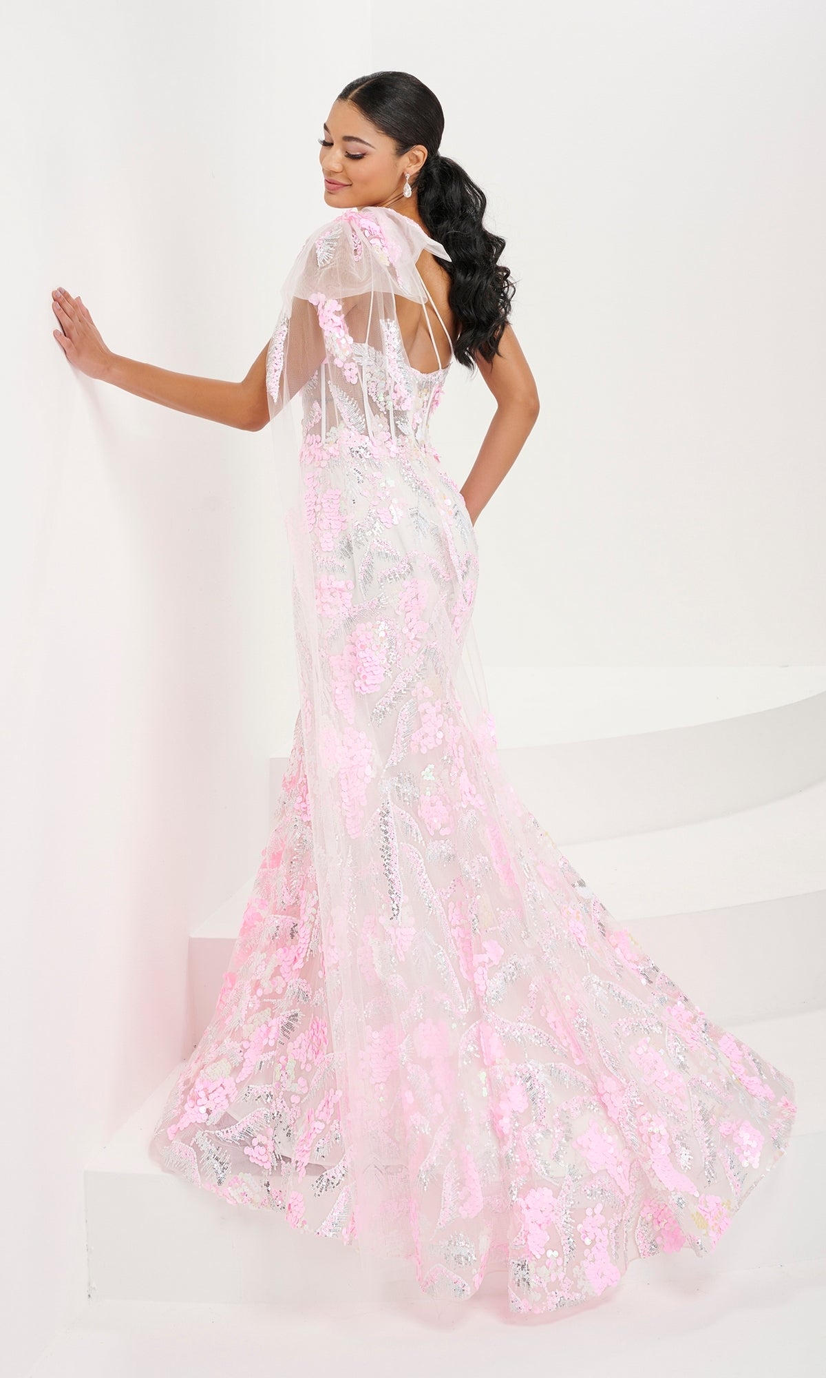 A woman poses on white stairs in the Tiffany One-Shoulder Sequin Sheer Prom Dress 16082, a fitted floor-length gown with a sheer bodice and pink floral embroidery, standing partly turned away and touching the wall.
